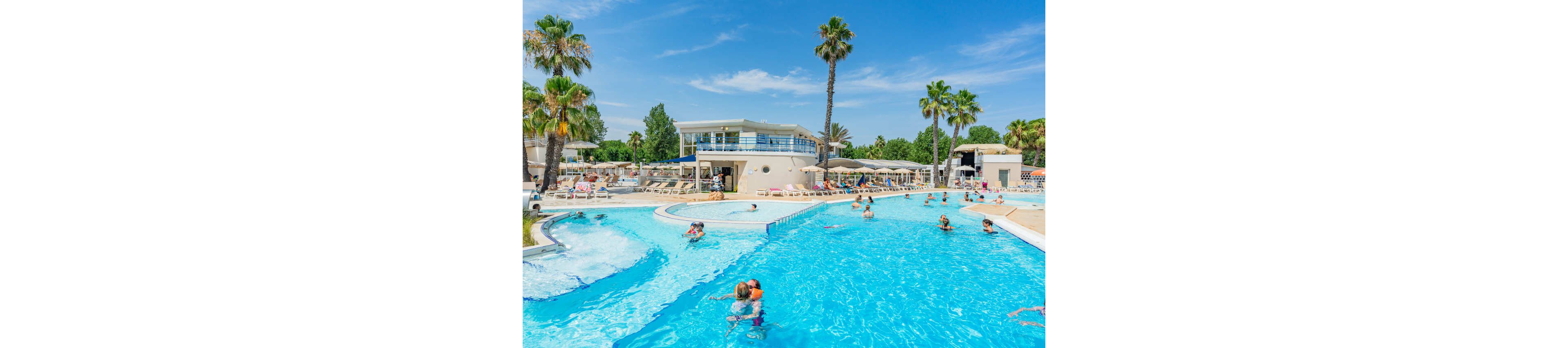 A large outdoor swimming pool is filled with families enjoying a sunny day at a resort. Children and adults play in the bright blue water while others relax on loungers around the pool. Palm trees and a modern white building with an upper deck overlook the scene. The clear sky and vibrant colors create a cheerful, vacation atmosphere.