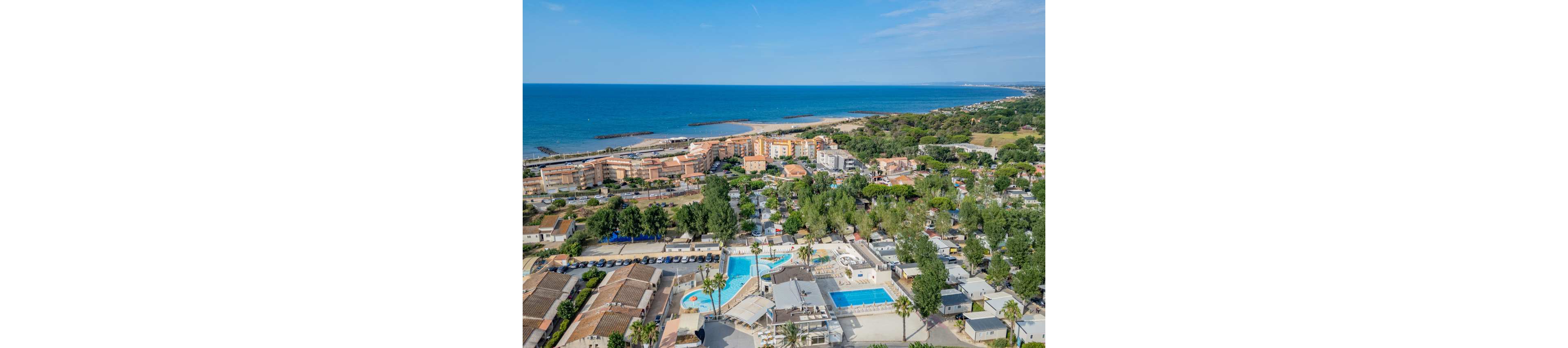 An aerial view shows a coastal resort town beside a wide sandy beach and calm blue sea. Residential buildings and holiday accommodations are clustered near several bright blue swimming pools. Green trees and landscaped areas are interspersed among the structures, creating a relaxed vacation atmosphere. The coastline stretches into the distance under a clear blue sky, suggesting a warm, sunny day. Cars are parked along the streets, indicating an active yet tranquil seaside destination.