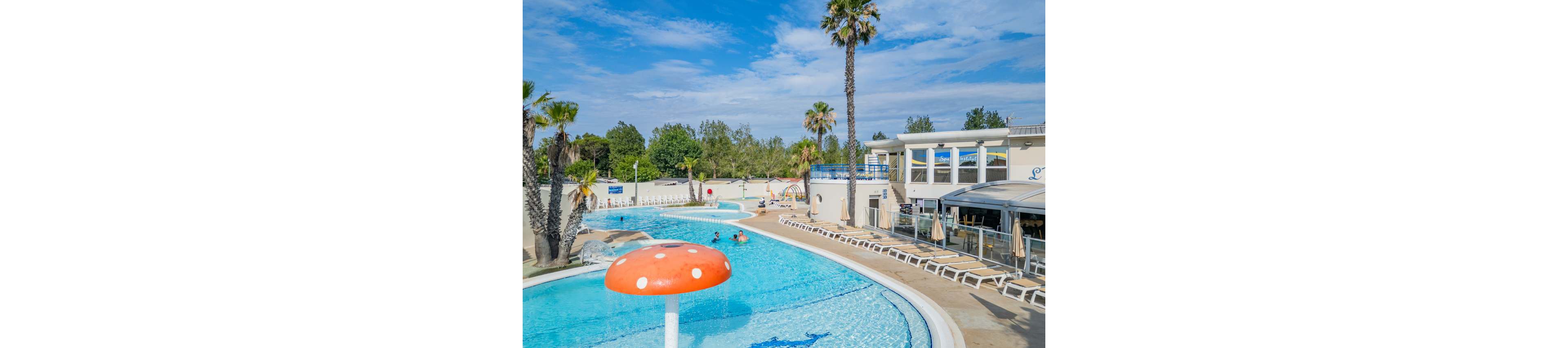 An outdoor resort-style swimming pool curves around a modern white building under a bright blue sky. A large red and white mushroom-shaped fountain stands in the shallow water near the foreground. Palm trees and greenery surround the pool area, while rows of white lounge chairs line the deck. A few people enjoy the water, creating a relaxed vacation atmosphere.