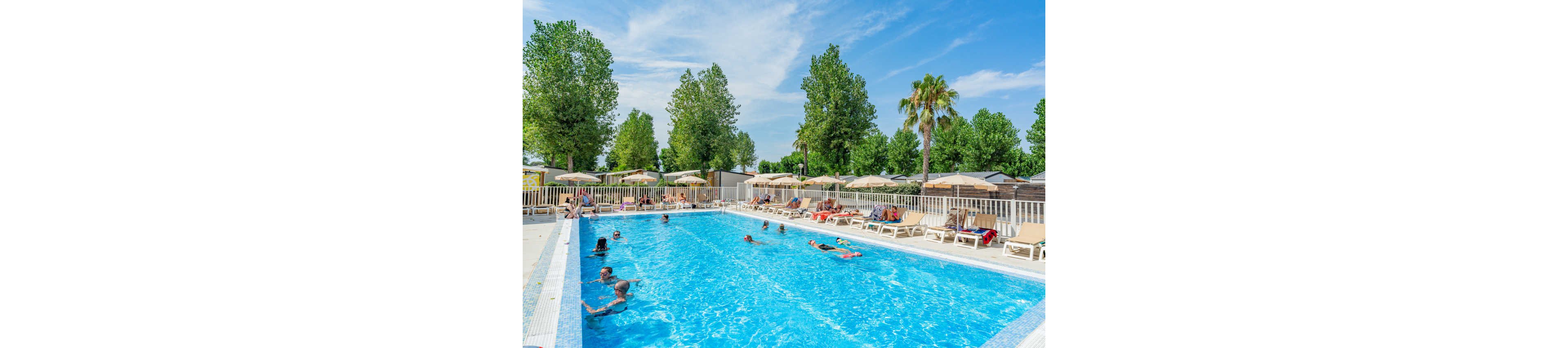 An outdoor swimming pool is filled with people enjoying a sunny day. Adults and children relax in the clear blue water while others lounge on sunbeds around the pool. Beige umbrellas line the deck, providing shade for guests. Tall green trees and a few palm trees surround the fenced pool area, creating a relaxed resort atmosphere.