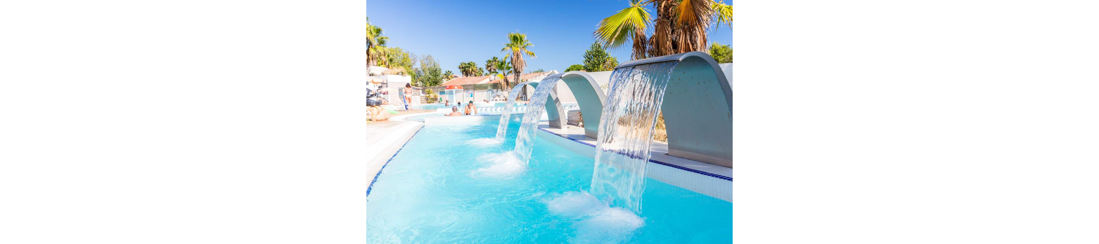 A bright resort swimming pool features multiple curved metal waterfalls pouring clear water into the turquoise pool. Palm trees and white buildings surround the area, creating a sunny vacation atmosphere. People relax in the background, enjoying the warm weather and refreshing water. The clear blue sky enhances the tropical, leisure-focused setting.
