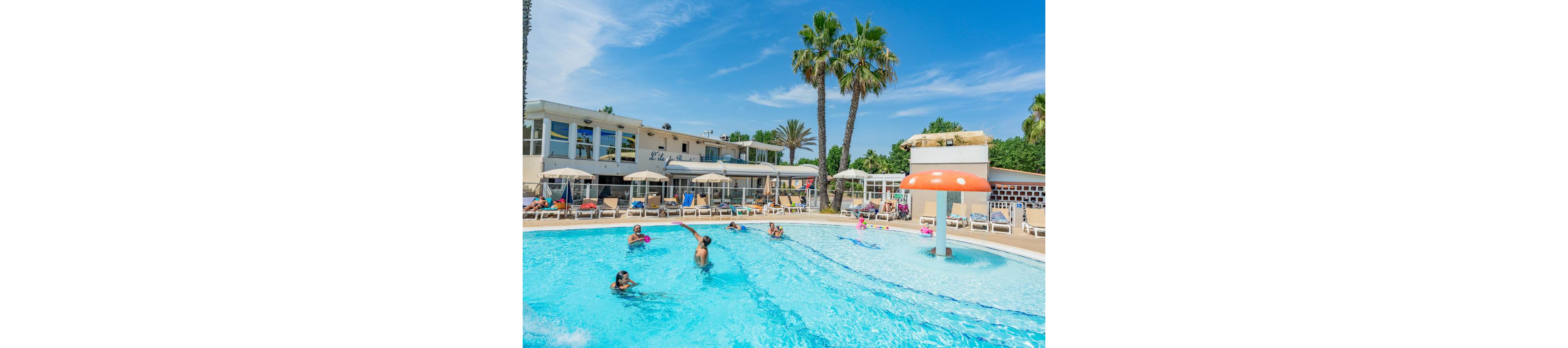 A bright outdoor swimming pool at a resort is filled with people enjoying the water. Children and adults play and swim under a clear blue sky, with lounge chairs and umbrellas lining the poolside. A playful mushroom-shaped fountain feature sprays water near the shallow end. Palm trees and a modern white building create a relaxed vacation atmosphere in the background.