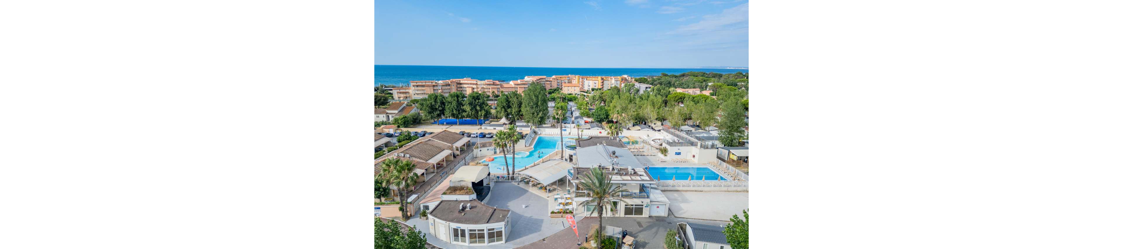 An elevated view shows a coastal resort complex featuring multiple swimming pools and sun terraces. Low-rise buildings and palm trees surround the pool area, creating a relaxed holiday atmosphere. In the background, residential blocks and lush greenery lead toward the shoreline and the open sea. Clear blue skies and calm water emphasize a bright, summery setting ideal for vacationers.