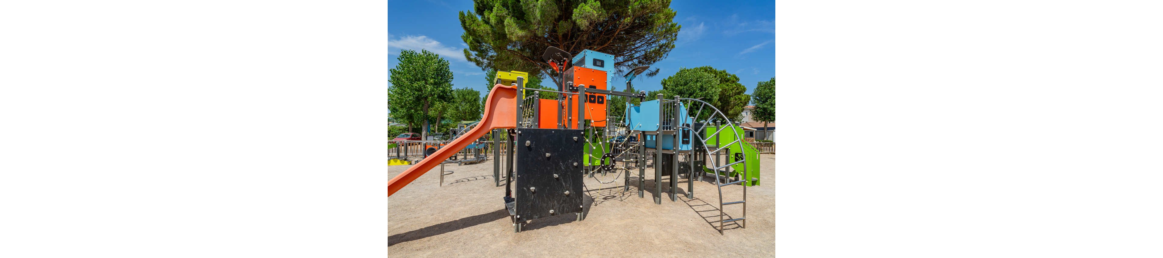 A brightly colored playground structure stands in an outdoor park. The equipment includes an orange slide, climbing walls, ladders, and metal arches designed for children. Trees and a clear blue sky frame the play area, suggesting a sunny day. The ground is covered with compacted sand or gravel, and no people are present in the scene.