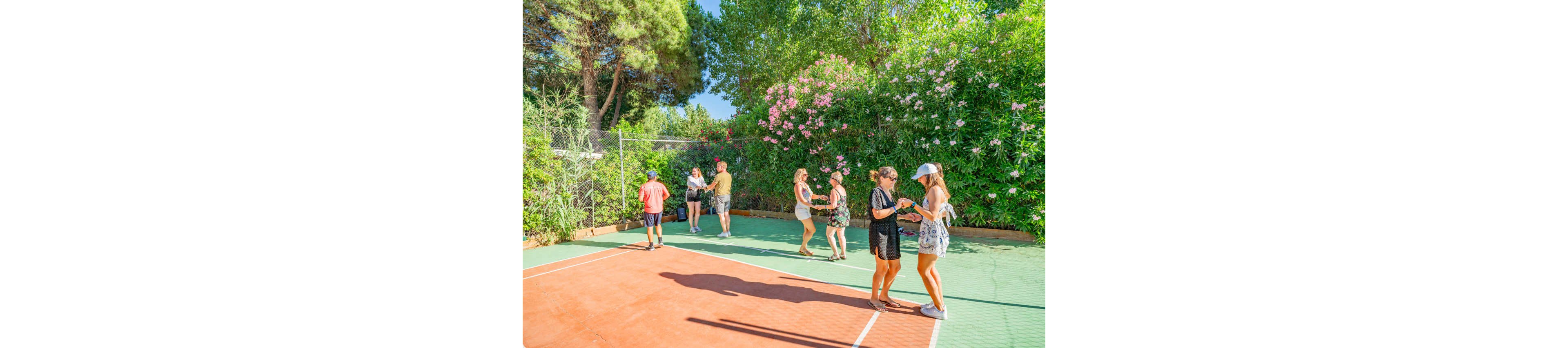 A group of people gather and socialize on an outdoor sports court surrounded by lush greenery. Several small groups stand chatting, some holding drinks or phones, while sunlight casts strong shadows on the ground. Tall trees and flowering bushes line the fence, creating a vibrant, summery backdrop. The relaxed scene suggests a casual meetup or vacation setting in warm weather.