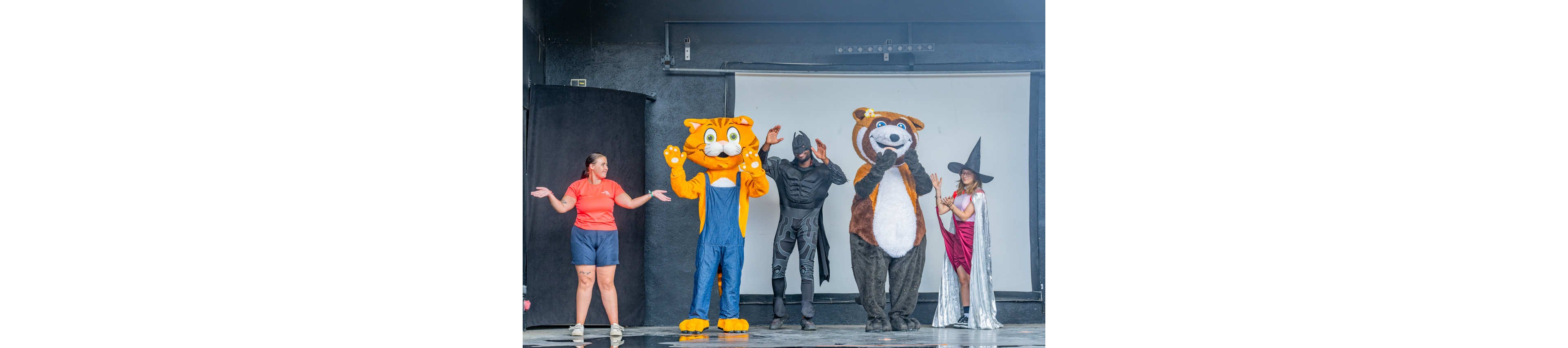 A group of performers stands on a stage dressed in colorful costumes. Two people wear large animal mascot suits, one resembling a cat and the other a bear, while another is dressed as a superhero and another as a witch. A casually dressed woman in shorts and a t-shirt joins them, all posing with playful gestures. The background is a simple dark stage with a white screen, suggesting a family-friendly show or event.