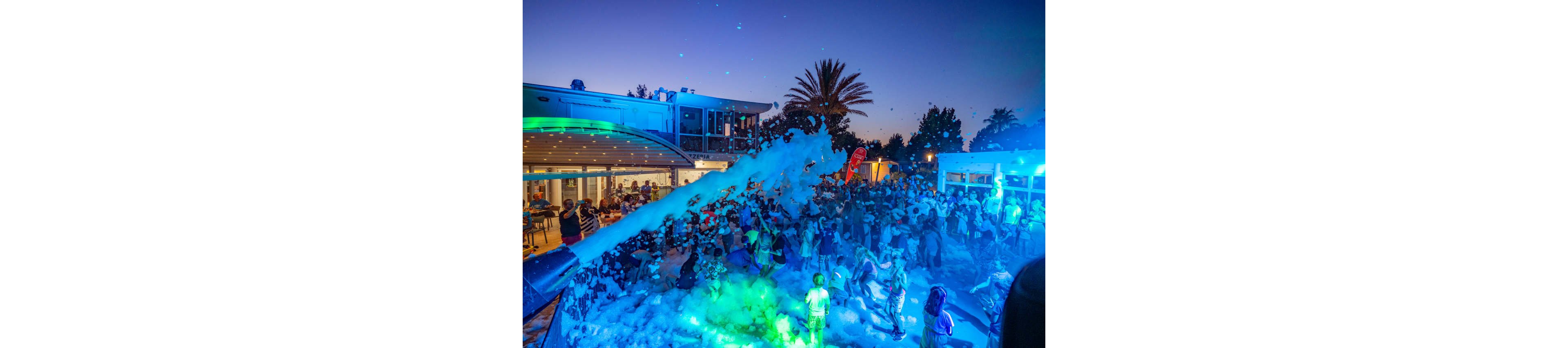 A large crowd of people gathers at an outdoor foam party in the evening. Thick foam covers the ground as it is sprayed from a large cannon in the foreground. Colorful blue and green lights illuminate the scene, contrasting with the warm glow from the building and restaurant area behind the crowd. Palm trees and a clear twilight sky frame the festive atmosphere, emphasizing a lively summer nightlife setting.