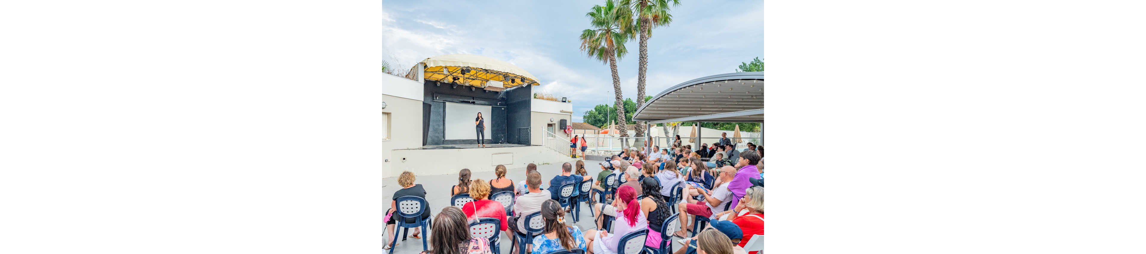 A woman stands on an outdoor stage speaking or performing in front of a seated audience. The crowd sits on plastic chairs arranged in rows, facing the small open-air theater. Palm trees and a partly cloudy sky create a relaxed, summery atmosphere. The scene suggests a casual show, presentation, or community event at a resort or recreational venue.
