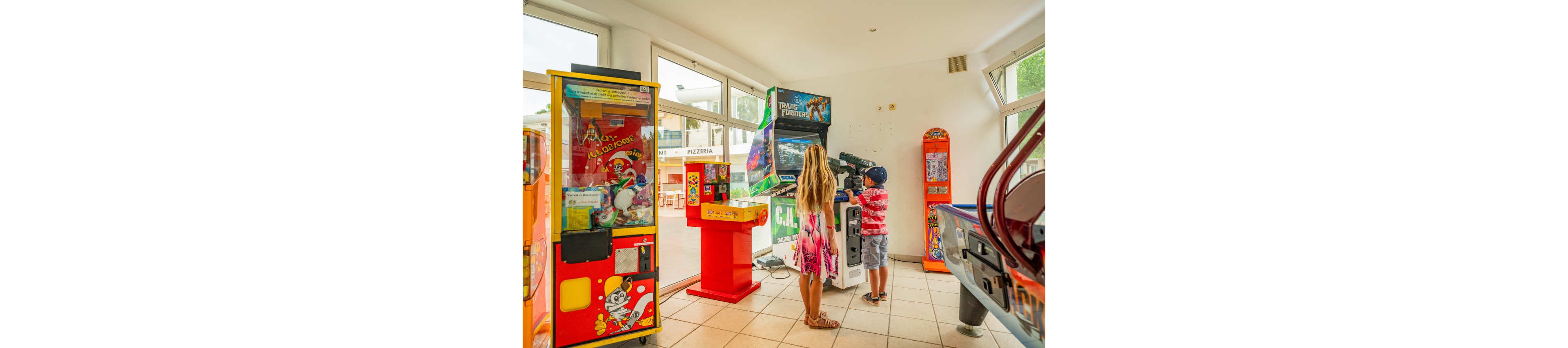 Two children stand at an arcade machine in a brightly lit game room. The girl in a pink dress and the boy in a striped shirt and cap focus on the screen together. Around them are various arcade cabinets, including a claw machine and a pinball-style game, with colorful graphics and characters. Large windows let in natural light and reveal a casual dining area outside.
