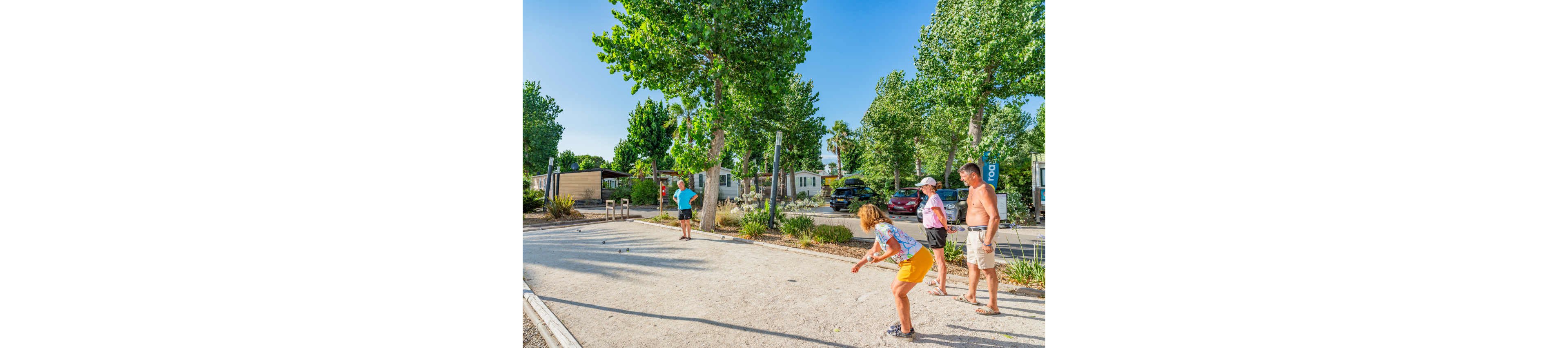 A group of adults play pétanque on a sandy court in a sunny outdoor campsite. Mobile homes, parked cars, and lush green trees surround the recreational area. One woman in the foreground is about to throw a boule while others watch and wait their turn. The bright daylight and casual summer clothing create a relaxed holiday atmosphere.
