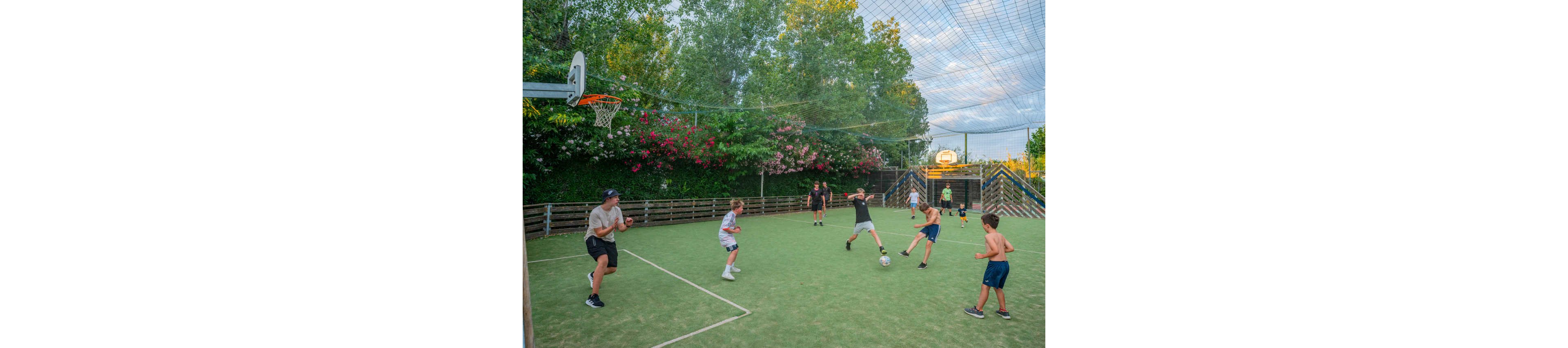 A group of children play soccer on a fenced outdoor sports court while an adult supervises. The court is covered with netting overhead and surrounded by lush green trees and flowering bushes. Some kids chase the ball while others watch or run across the field, creating a lively scene. Basketball hoops are visible at each end, highlighting the multi-sport nature of the space.