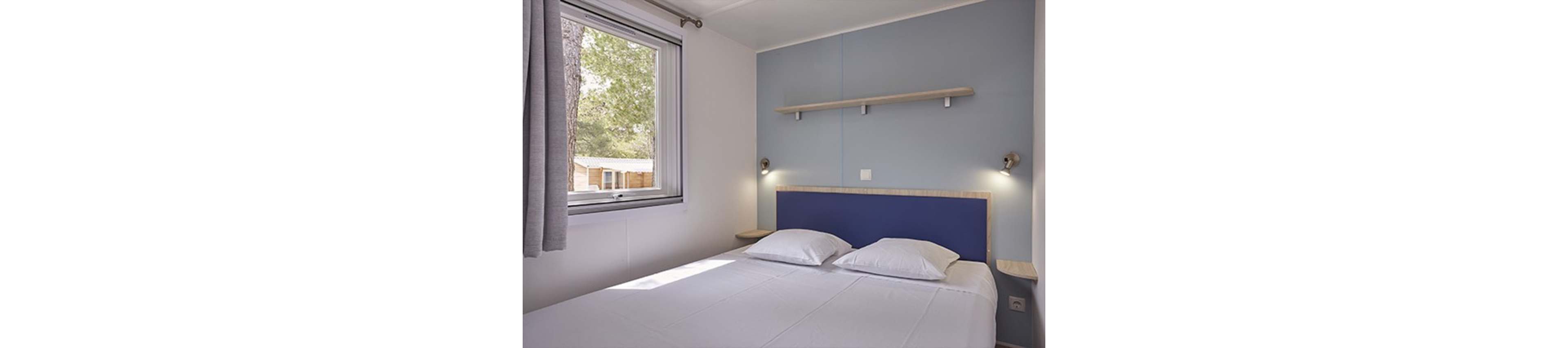 A small, minimalist bedroom features a double bed with a blue headboard and two white pillows. The room has light-colored walls and a soft blue accent wall with a simple wooden shelf above the bed. Natural light enters through a large window with curtains, revealing trees and another cabin outside. The overall atmosphere is calm, clean, and modern, ideal for a cozy stay.