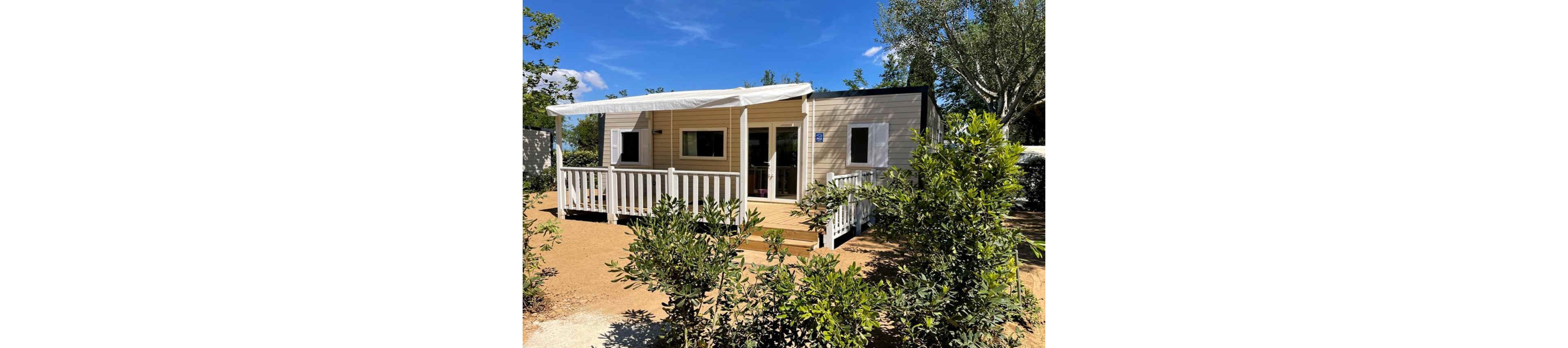 A modern mobile home with a white-railed veranda stands in a sunny outdoor setting. The structure features light-colored siding, multiple windows, and a covered terrace area. Green shrubs and small trees surround the home, adding natural privacy and a relaxed atmosphere. Clear blue sky and bright daylight suggest a warm, pleasant climate ideal for vacation or camping stays.