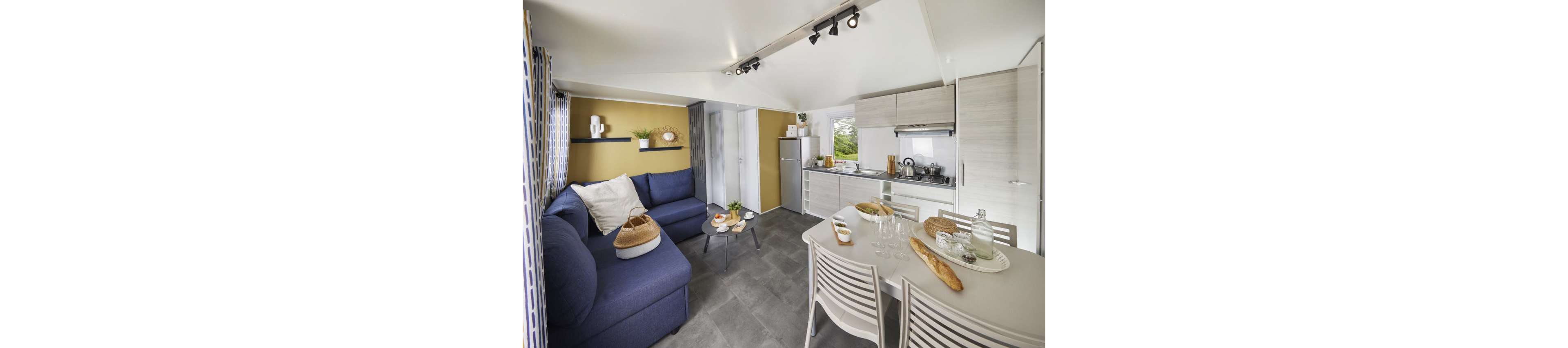 A modern open-plan interior combines a compact kitchen and a cozy living area. The space features a blue sectional sofa with cushions, a small coffee table, and a dining table set with glasses, plates, and bread. Light-colored cabinetry, a refrigerator, and a cooktop line the kitchen wall, while decorative shelves and plants add warmth. Track lighting and large windows brighten the room, highlighting the neutral tones and contemporary design.