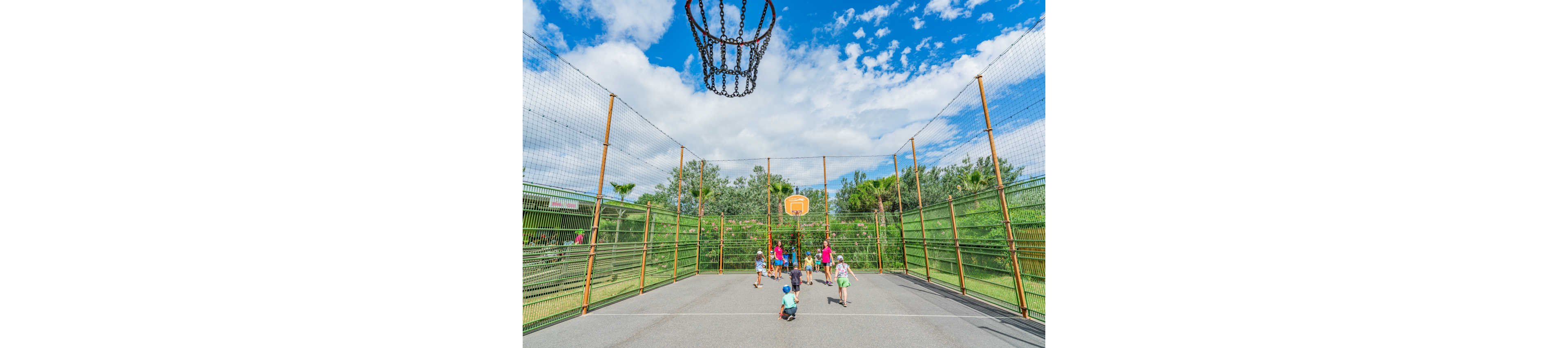 A group of children play basketball on an outdoor court surrounded by tall metal fencing. The scene is set in a green park with trees and bushes in the background. A basketball hoop is visible at the far end of the court, with another hoop and chain net seen from above in the foreground. The bright daylight and blue sky with scattered clouds create a cheerful, energetic atmosphere.