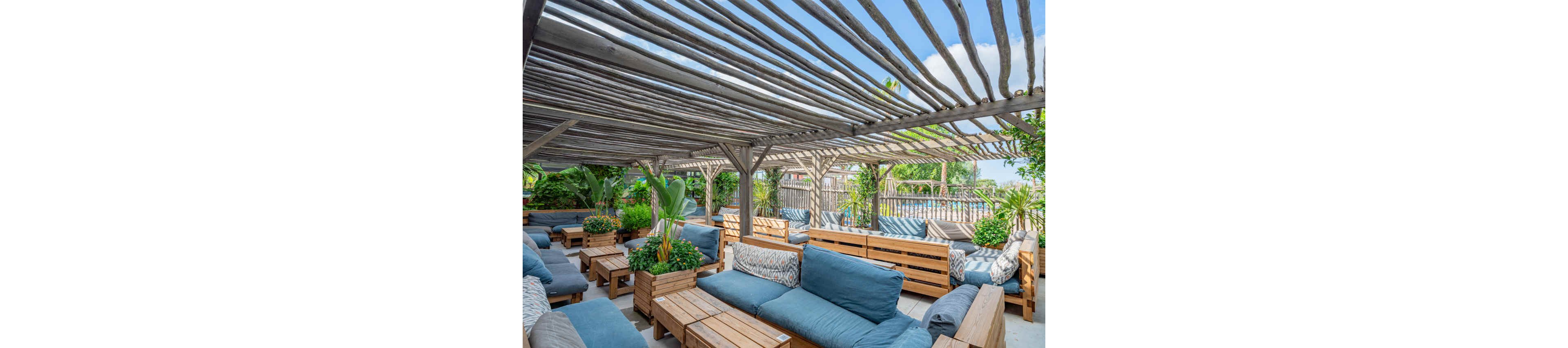An inviting outdoor lounge area features multiple wooden sofas with blue cushions arranged under a slatted wooden pergola. Potted tropical plants and greenery are placed throughout the space, adding a lush, natural feel. Decorative patterned pillows enhance the cozy seating arrangement. In the background, a pool and additional seating can be seen under a bright, partly cloudy sky.