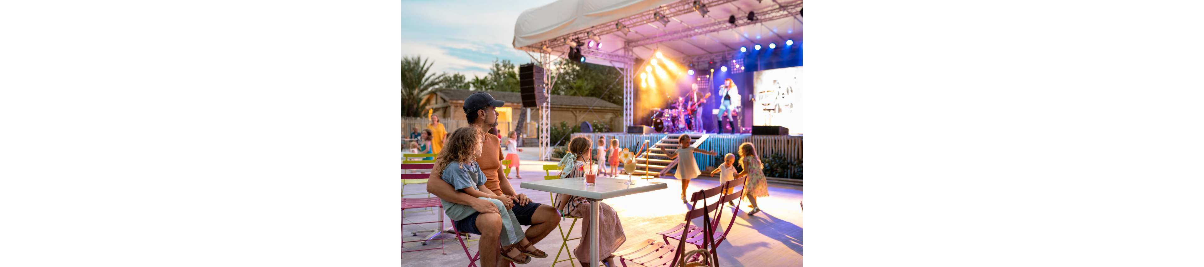 A father sits with his young daughter at a colorful outdoor venue while watching a live band perform on stage. Children play and dance near the tables and in front of the lit stage. Bright stage lights and a large screen highlight the musicians in the background. The scene captures a relaxed summer evening atmosphere with families enjoying entertainment together.