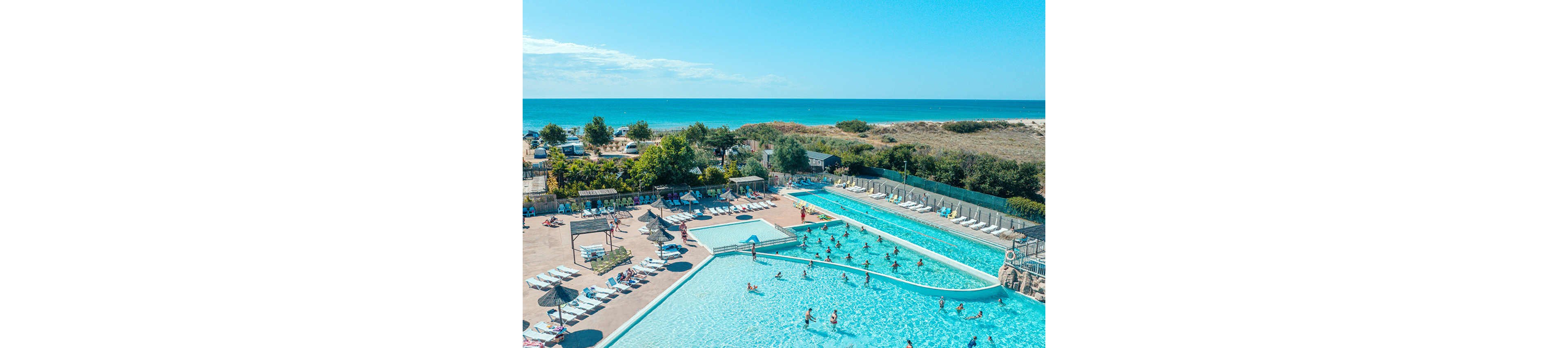A large outdoor swimming pool complex is shown near a sandy coastline with blue ocean in the background. Multiple pools are filled with people swimming and playing in the water. Sun loungers and parasols surround the pool area, with guests relaxing under the sun. Trees, dunes, and parked vehicles separate the resort from the beach, creating a relaxed holiday atmosphere.