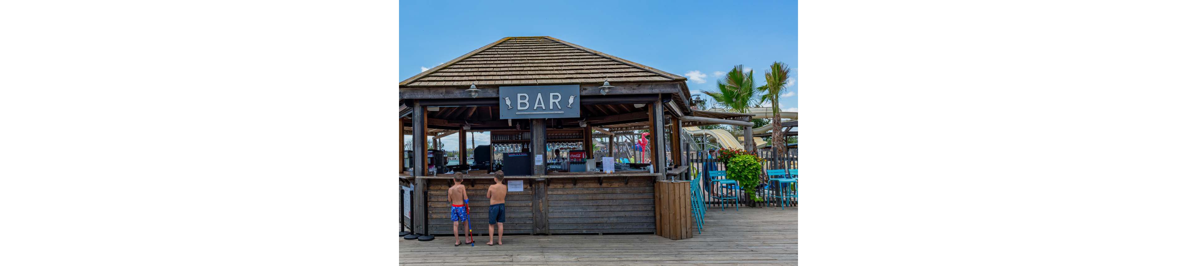 A wooden open-air bar stands on a boardwalk near the beach. Two young boys in swim trunks wait at the counter, suggesting a casual vacation setting. Palm trees, outdoor seating, and water slides are visible in the background under a clear blue sky. A large sign reading "BAR" hangs above the counter, emphasizing the relaxed, seaside refreshment spot.
