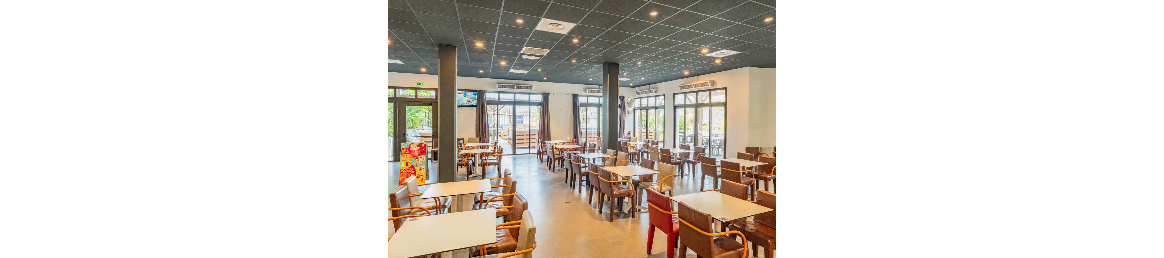 Spacious cafeteria interior with rows of tables and wooden chairs is shown empty and ready for guests. Large windows and glass doors line the walls, letting in natural light and views of the outdoor seating area. The ceiling features a grid of recessed lights and ventilation panels, while neutral floors and walls create a clean, modern look. A colorful refrigerated display cabinet stands near the entrance, adding a pop of color to the scene.