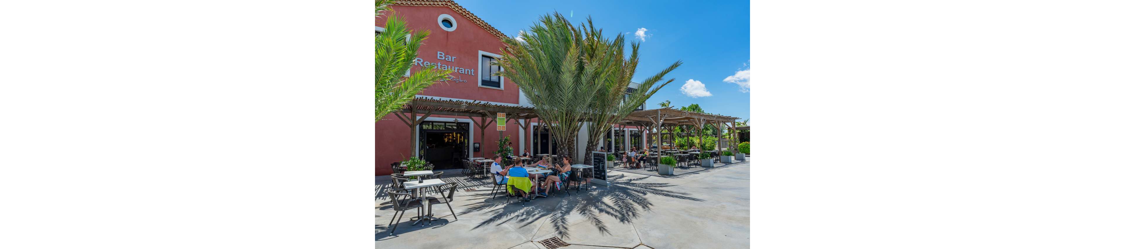 People sit at tables on the outdoor terrace of a bar restaurant under wooden pergolas and tall palm trees. The building has a warm reddish facade with large open doorways leading inside. Bright sunlight and blue sky create a relaxed holiday atmosphere. Diners appear to be enjoying food and drinks in a casual, open-air setting.