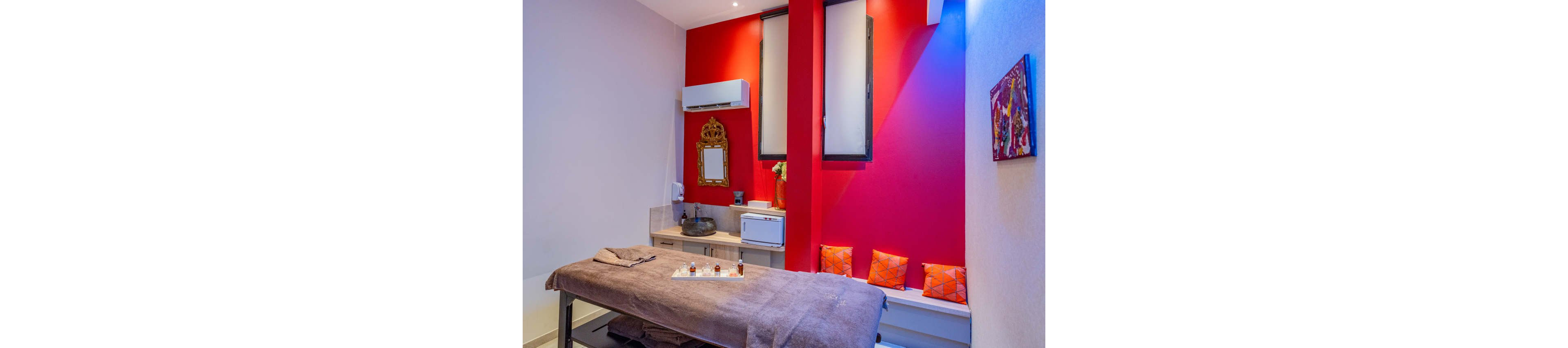 A neatly arranged spa treatment room features a massage table covered with brown linens and small bottles of oils on a tray. The space has bold red accent walls contrasted with light neutral tones and soft lighting. Cushions with geometric orange patterns sit on a bench beneath tall windows. A decorative framed mirror, countertop basin, and compact appliances complete the serene wellness setting.