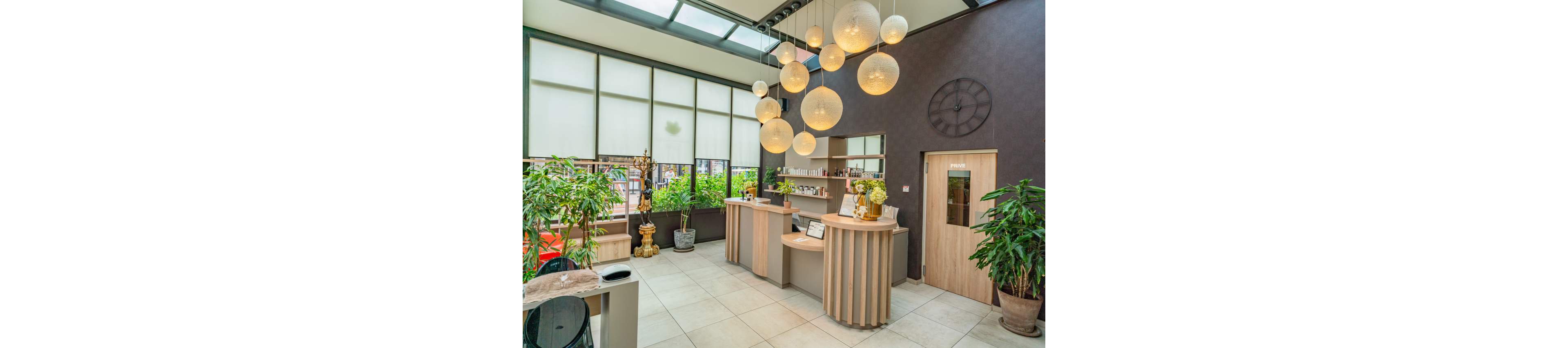 Contemporary spa reception area featuring curved wooden desks and a wall of product shelves. Large spherical pendant lights hang from the ceiling, illuminating the bright space. Tall windows with translucent blinds line the back wall, with green plants adding a natural touch. A wooden door with a small glass window and a large wall clock complete the clean, modern interior.