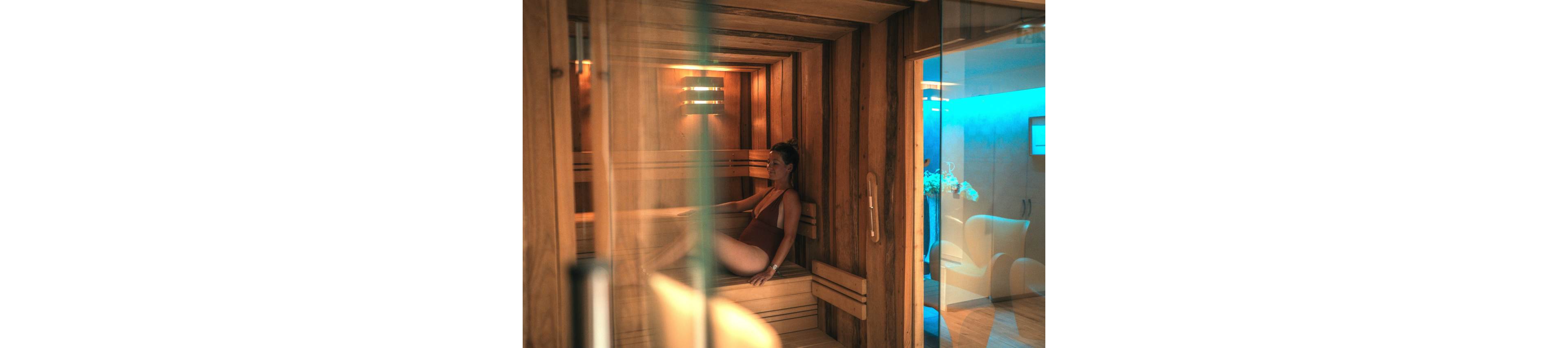 A woman in a swimsuit relaxes inside a warm wooden sauna. She is seated on a bench with her back against the wall, enjoying a quiet moment. Soft lighting from a wall fixture enhances the cozy atmosphere of the sauna cabin. Through the glass door, a modern spa lounge with chairs and blue ambient light is visible in the background.