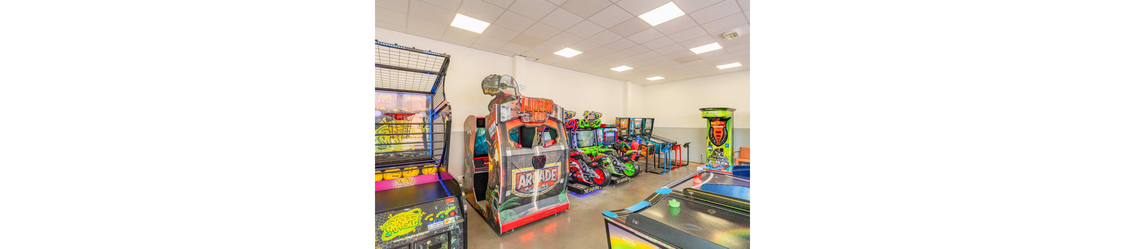 A bright indoor arcade room features a variety of colorful gaming machines lined up against a white wall. Large stand-up arcade cabinets, including a racing-style machine and motorcycle games, dominate the scene. Additional pinball machines and an air hockey table are visible in the foreground. The space is well-lit by a grid of ceiling lights, creating a clean and inviting entertainment area.