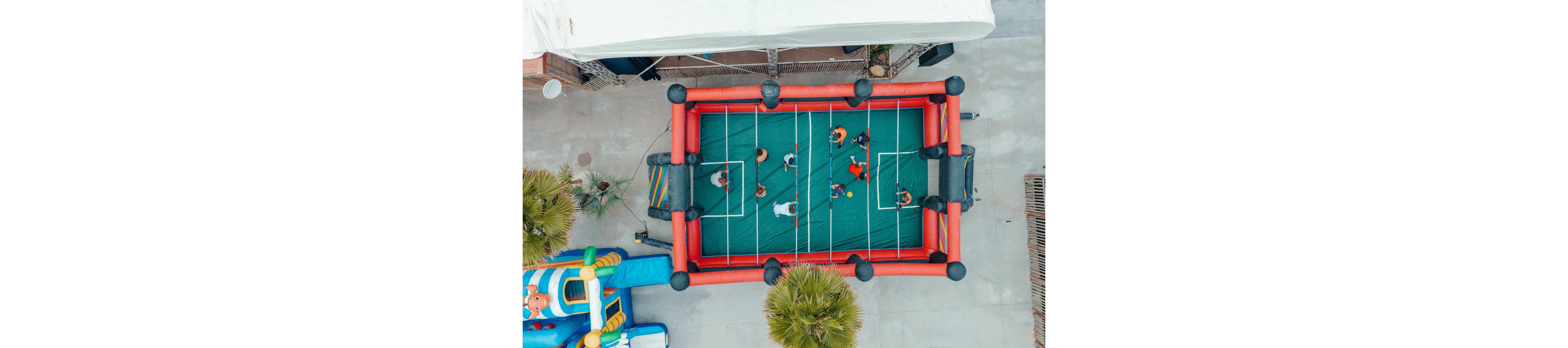 A group of people play on a large inflatable human foosball court viewed from above. The green playing field is surrounded by red and black inflatable borders with white lines marking the goals and lanes. Players are spaced across the field, each holding onto horizontal bars as they face their opponents. Nearby, an inflatable play structure and palm trees suggest a fun outdoor event or party setting.