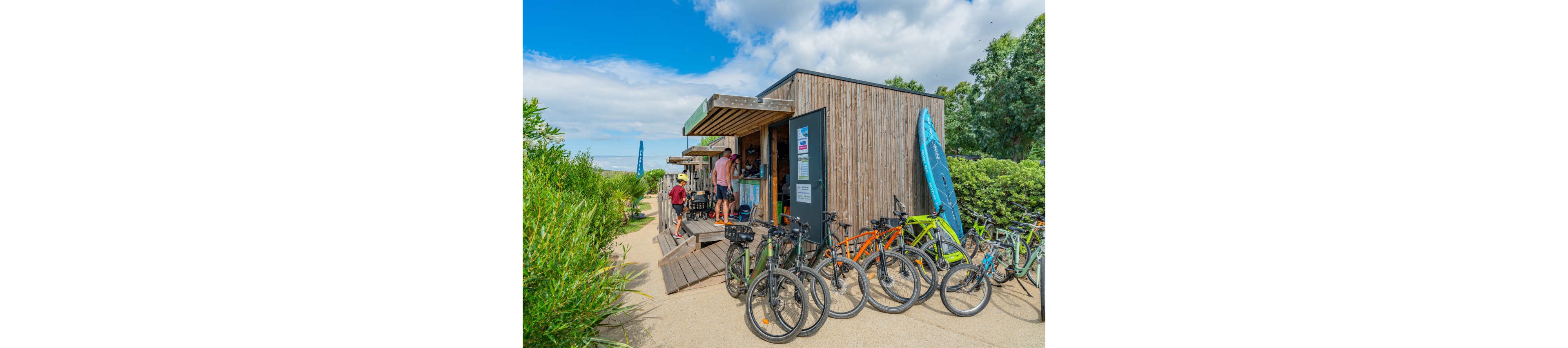 A small wooden bike rental shop stands beside a sandy path lined with greenery. Several bicycles are parked in front, while people gather at the counter to rent equipment. A bright blue kayak is stored upright against the side of the building, suggesting additional water sports activities. The scene is set under a partly cloudy blue sky, creating a relaxed, summery atmosphere.