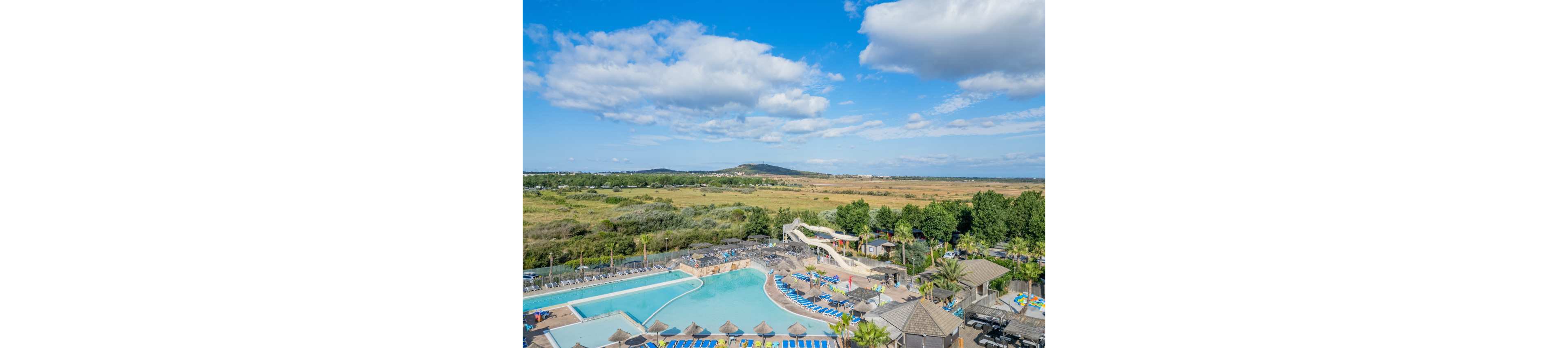 A large resort complex features multiple outdoor swimming pools with clear blue water. Numerous sun loungers and parasols surround the pools, and a tall water slide curves into one of the basins. The resort is bordered by lush green trees and opens onto expansive fields and distant hills under a bright blue sky. Puffy white clouds add depth to the wide, sunny landscape view.