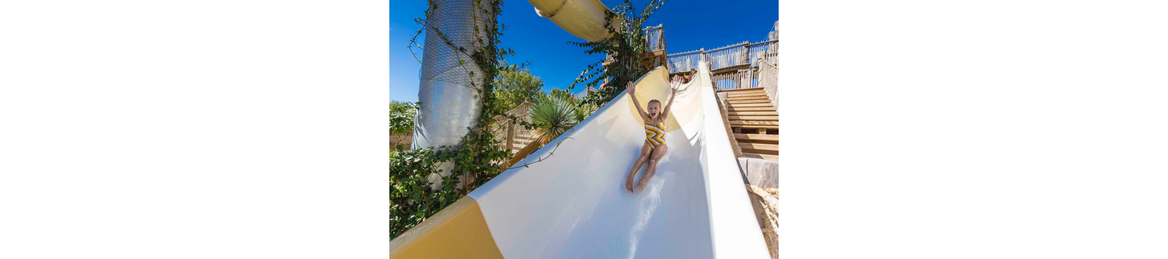 A young child in a patterned swimsuit rides down a large outdoor waterslide with arms raised in excitement. The slide is white with yellow sections and surrounded by greenery and wooden structures. Bright blue sky and sunlight create a vivid, summery atmosphere. The scene captures the fun and energy of a day at a water park or resort.