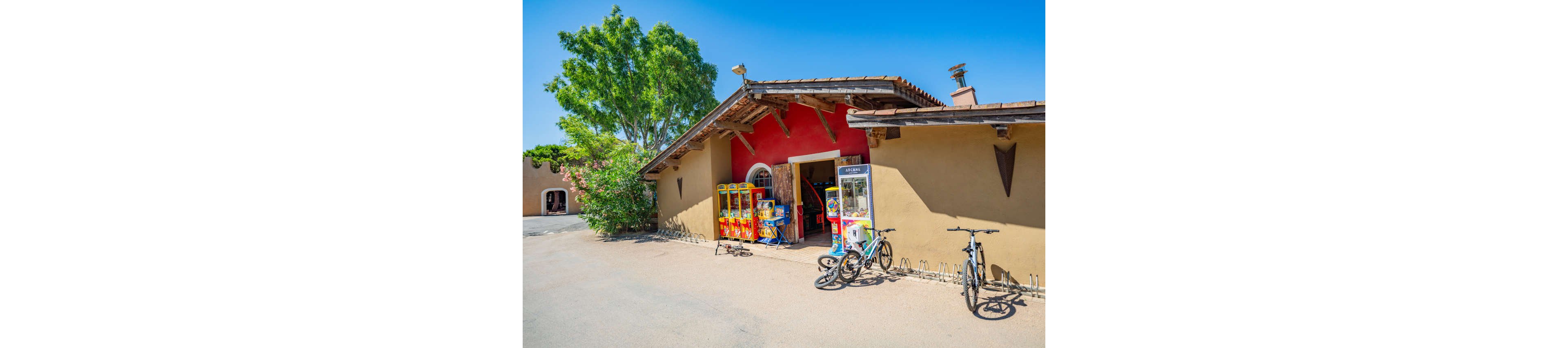 A rustic building with a red accent wall features several colorful arcade and claw machines near its open entrance. Two bicycles are parked in front of the structure, suggesting visitors have arrived by bike. The scene is set on a bright, sunny day with clear blue skies and a large green tree providing contrast to the tan stucco walls. The overall atmosphere feels relaxed and playful in an outdoor recreational area.