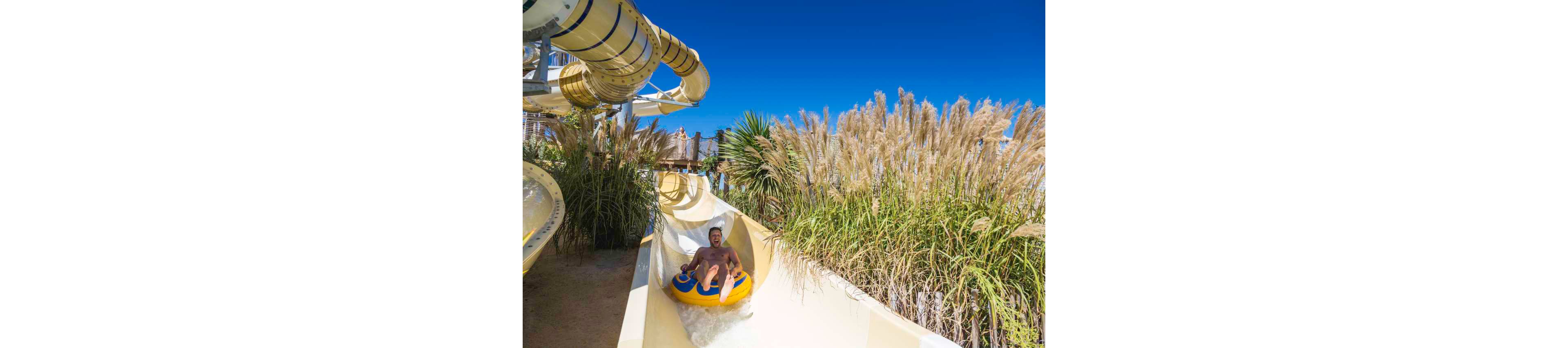A man rides a yellow inflatable tube down a curved water slide. Tall ornamental grasses line the slide, creating a lush, natural atmosphere. The sky above is clear and intensely blue, suggesting a bright sunny day. The scene captures a fun, energetic moment at an outdoor water park.