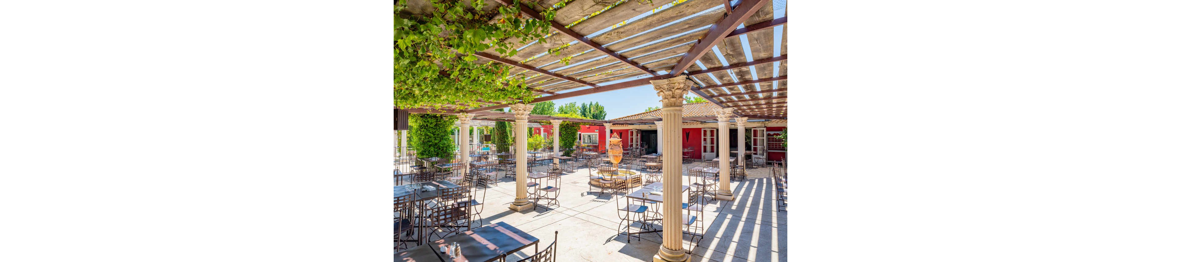 An open-air restaurant terrace features numerous metal tables and chairs arranged beneath a wooden pergola. Sunlight filters through the slatted roof, partially covered with green vines, casting patterned shadows on the stone floor. Classical-style columns support the structure, framing views of colorful building facades and large windows in the background. The bright, clear sky and lush greenery create a relaxed Mediterranean ambiance ideal for outdoor dining.