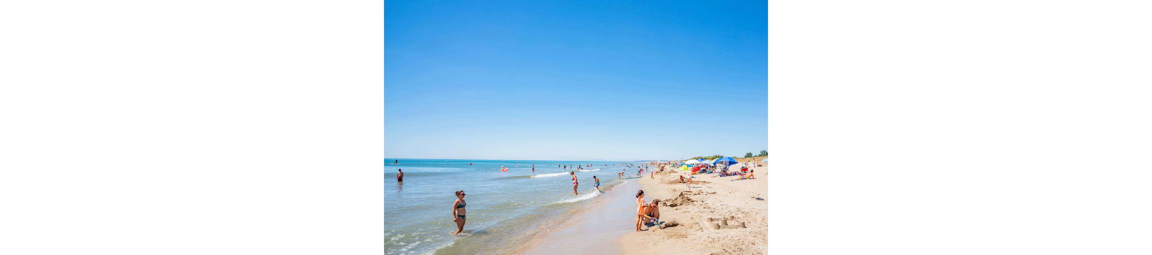 People relax and play along a sunny sandy beach beside calm blue water. Swimmers wade in the shallow shoreline while others stand or walk on the wet sand. Colorful umbrellas and towels are set up further up the beach, suggesting a busy summer day. The clear blue sky and bright sunlight create a warm, cheerful atmosphere.