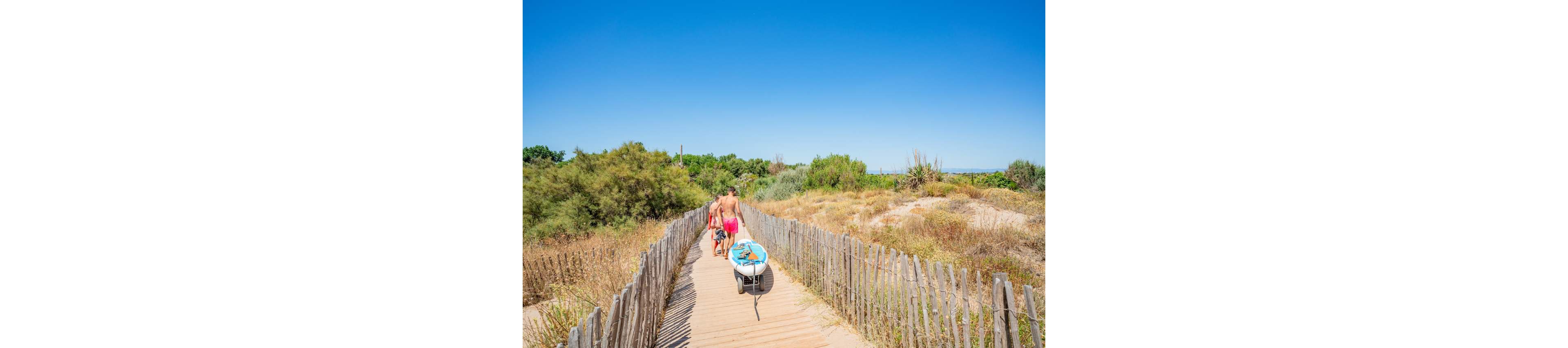 A man and child walk along a wooden boardwalk toward the beach on a bright sunny day. The man pulls a small cart loaded with beach gear, including a blue inflatable ring. Weathered wooden fences line the path, with dry grasses and shrubs on either side. The sky is clear and intensely blue, creating a warm, summery atmosphere.