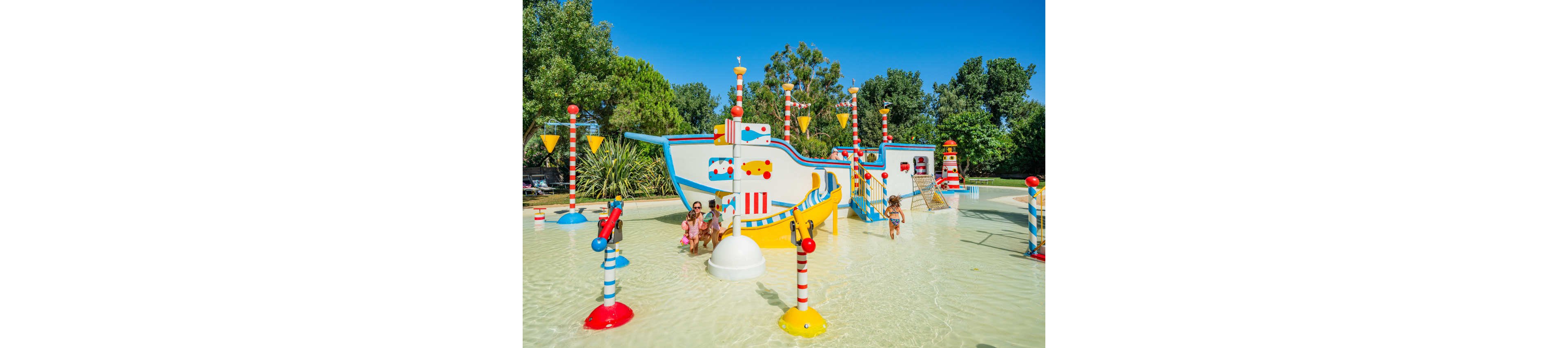 Young children play on a brightly colored pirate-themed water playground shaped like a ship. The shallow splash pool is surrounded by whimsical striped poles and interactive water features. Lush green trees and clear blue sky create a sunny outdoor setting. The scene feels cheerful and playful, emphasizing family fun and summertime recreation.
