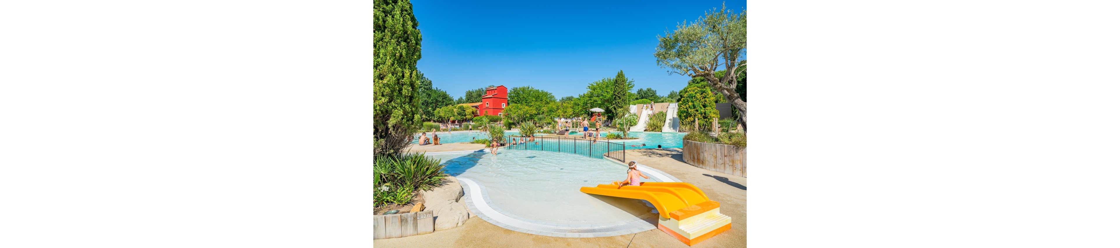 A shallow pool with a small yellow slide is filled with children playing under the sun. Families relax and swim in the larger pools and water features in the background. Lush greenery, trees, and landscaped plants surround the aquatic area, creating a resort-like atmosphere. A bright red building and clear blue sky complete the cheerful summer scene.