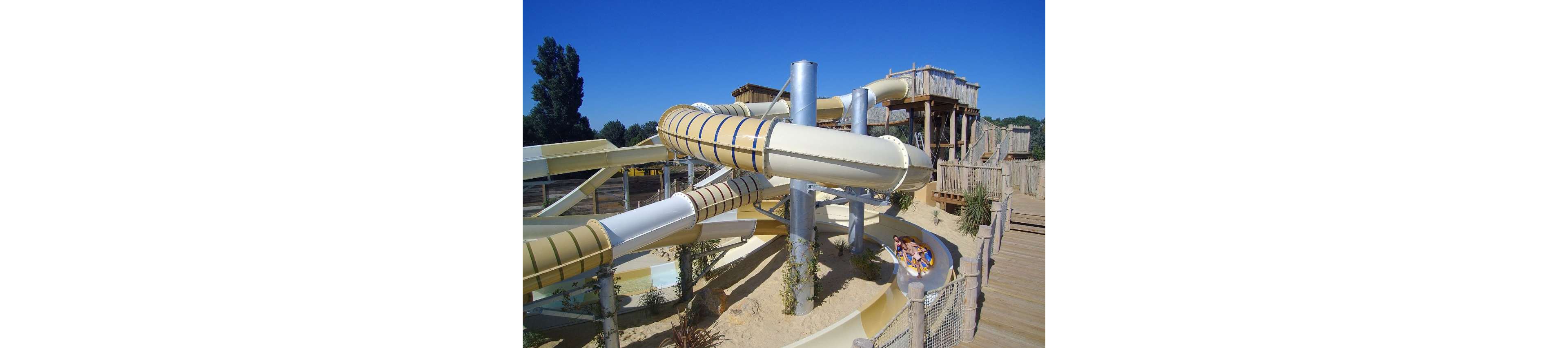 A large twisting waterslide winds around metal supports in an outdoor water park. Several people ride together in a raft as they exit one of the enclosed slide tubes. Wooden platforms, railings, and walkways surround the slide structure, creating a rustic adventure setting. The scene is bright and sunny, emphasizing leisure and summertime fun.