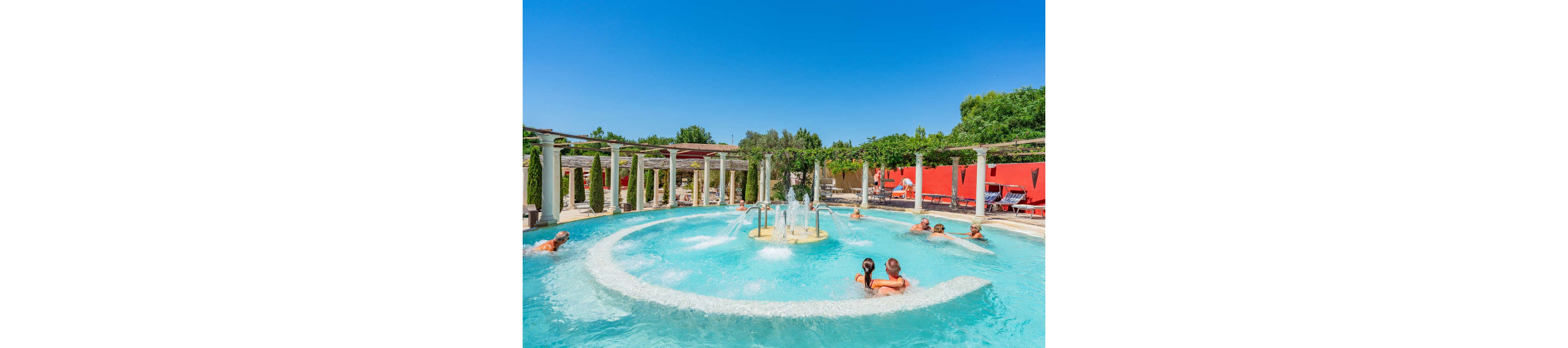 An outdoor circular pool features multiple water jets and fountains under a clear blue sky. Several adults relax in the warm water around the central fountain area. The pool is surrounded by white columns, greenery, and red walls that add vibrant color to the scene. Sun loungers and shaded pergolas suggest a comfortable resort or spa environment.