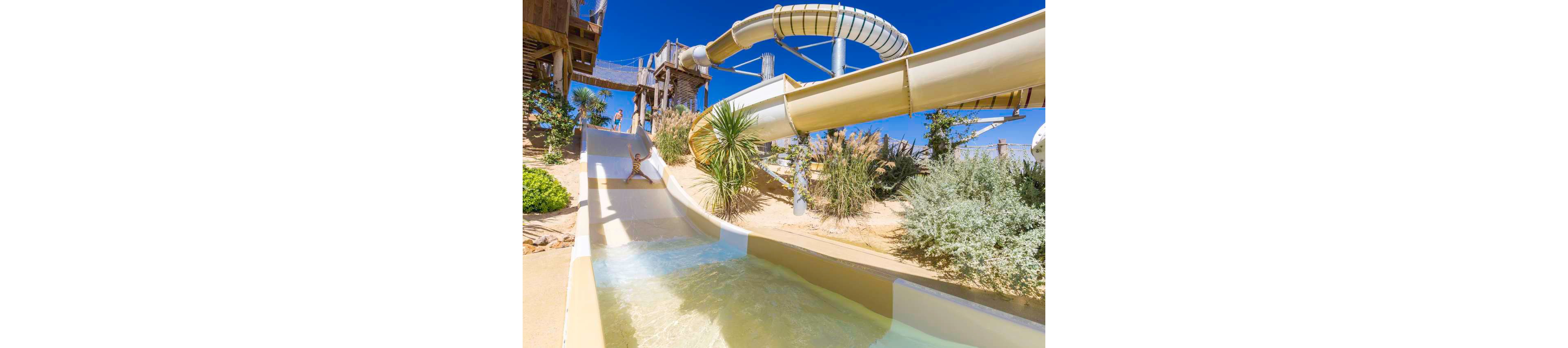 A young child rides down a large open water slide at an outdoor water park. The slide curves through a sandy, landscaped area with palm trees and ornamental grasses. Wooden structures and elevated walkways frame the attraction under a clear blue sky. Bright sunlight and vivid colors create a fun, summery atmosphere.