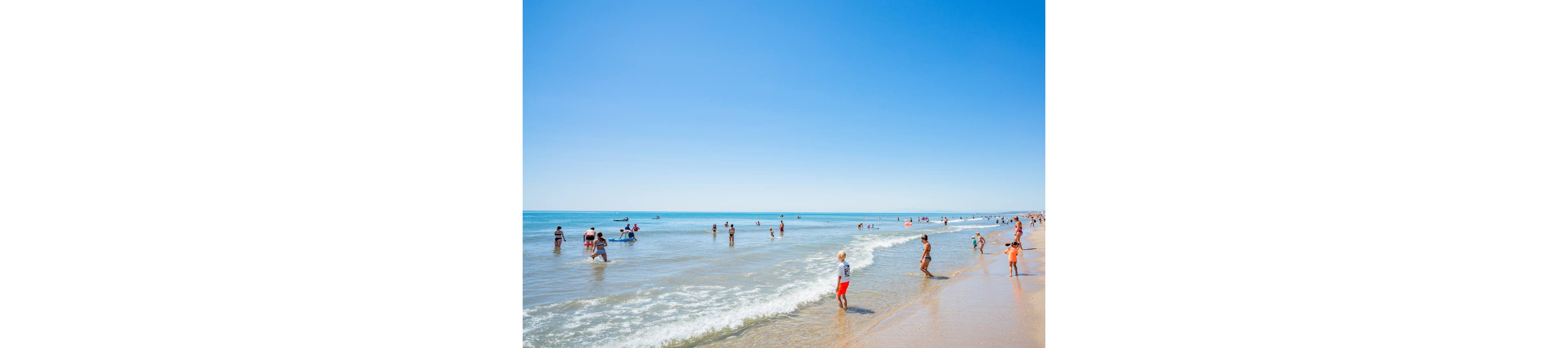 People of various ages play and relax along a wide sandy beach on a bright sunny day. Gentle waves roll onto the shore as children wade in the shallow water and adults stand or walk near the surf. The clear blue sky and calm sea create a peaceful, summery atmosphere. Colorful swimwear and beach gear add lively accents to the expansive coastal scene.