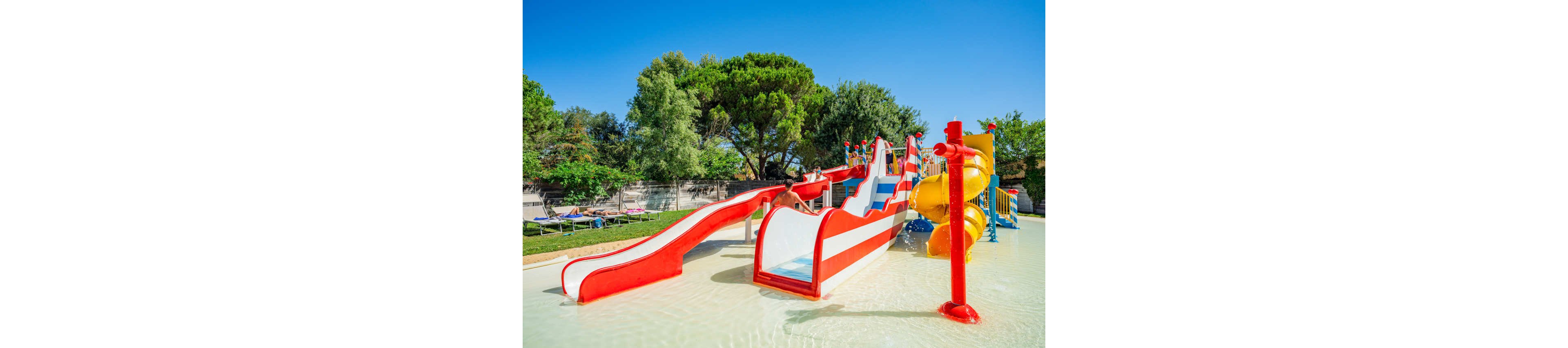 A bright red and white water slide structure stands in a shallow splash pool at an outdoor water park. Multiple slides and climbing sections are visible, including a twisting yellow tube slide. The area is surrounded by green trees, grass, and lounge chairs under a clear blue sky. The scene suggests a fun, family-friendly recreational space for children during warm weather.