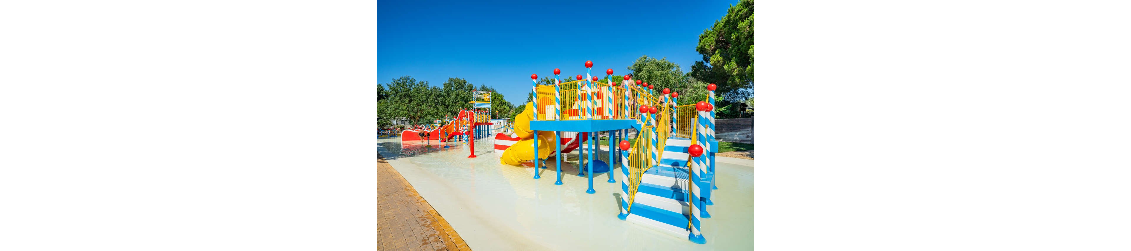 A brightly colored water playground structure stands in shallow water under a clear blue sky. The play area features striped stairs, platforms, and slides in blue, yellow, red, and white. Surrounding trees and a paved walkway frame the aquatic play zone, suggesting a family-friendly park or resort. The scene appears sunny and inviting, emphasizing fun and outdoor recreation for children.