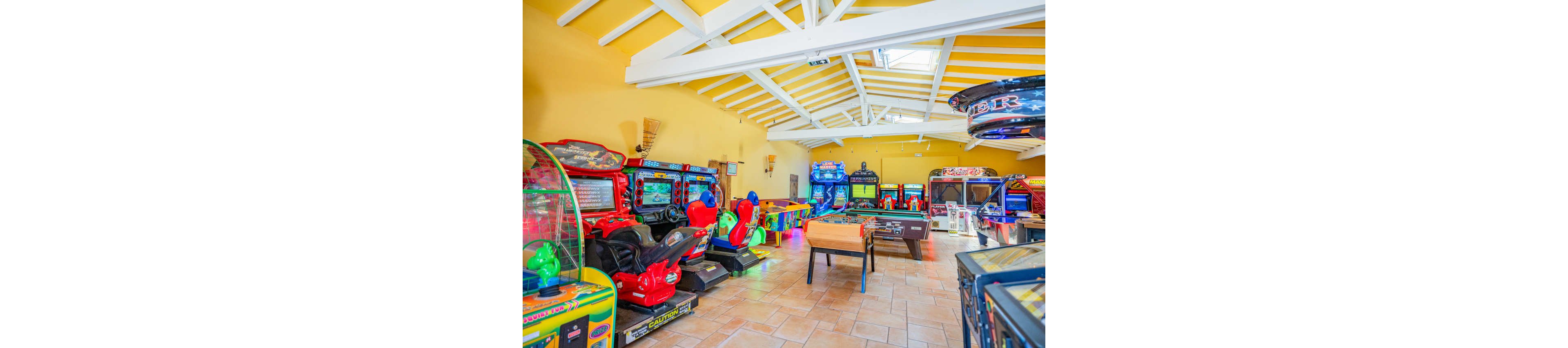 Brightly lit indoor arcade filled with various gaming machines and activities. Racing simulators, pinball machines, and other electronic games line the walls under a high white-beamed ceiling. A foosball table and air hockey table sit in the center of the tiled floor, inviting group play. Vibrant neon lights and colorful cabinets create an energetic, family-friendly entertainment atmosphere.