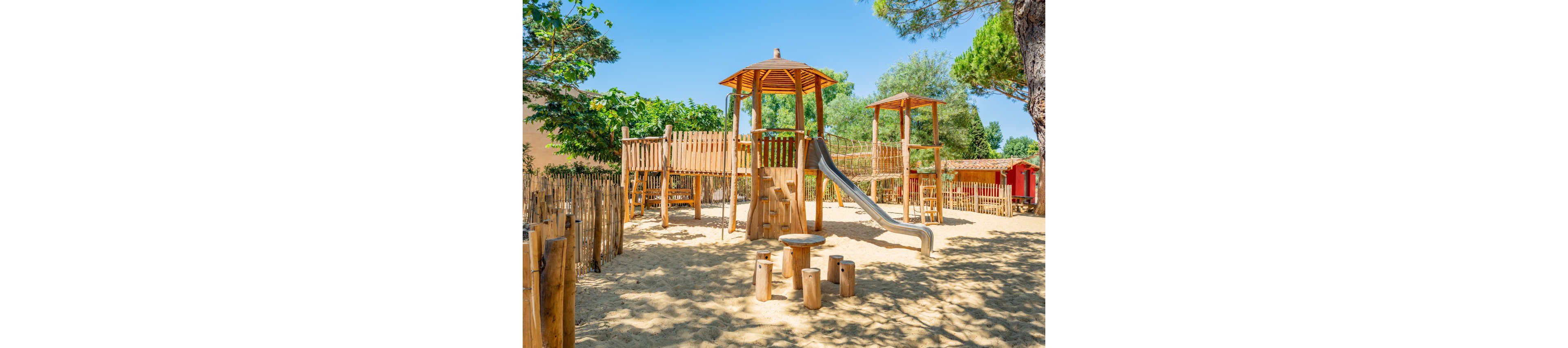 An outdoor wooden playground stands on a sandy surface under bright sunlight. The structure features towers with roofs, connecting walkways, and a metal slide. In the foreground, there is a small circular table with wooden stump seats. Trees, wooden fencing, and a red shed or cabin surround the play area, creating a natural and inviting setting.