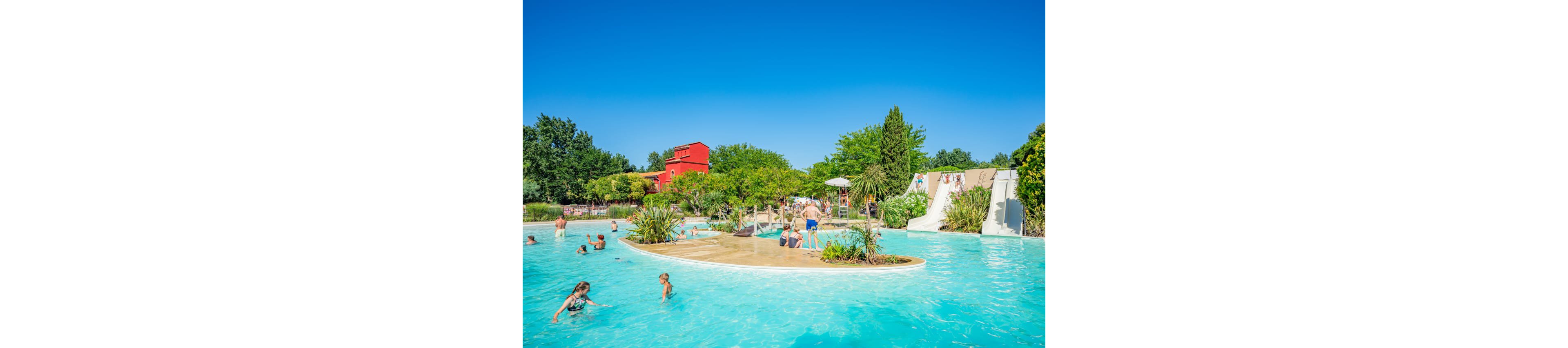 An outdoor swimming pool is filled with families and children enjoying a sunny day. People swim, wade, and play in the clear blue water around a planted island. A waterslide and small waterfall feature are visible in the background, adding to the fun atmosphere. Lush greenery and a bright red building surround the pool under a clear blue sky.