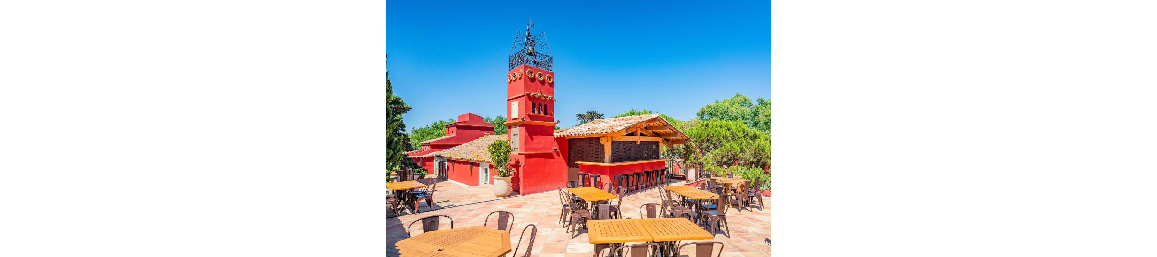 An open-air terrace bar features wooden tables and metal chairs under a clear blue sky. Bright red stucco buildings with tiled roofs and a decorative tower create a Mediterranean-style setting. The bar counter is shaded by a wooden pergola, and lush green trees surround the patio. The scene suggests a relaxed daytime atmosphere ideal for outdoor dining or social gatherings.